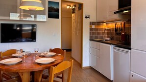 Cocina moderna con gabinetes blancos, horno integrado y campana extractora de acero inoxidable. Hay un comedor con mesa y sillas de madera, y un televisor de pantalla plana montado en la pared.