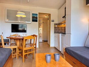 Kompakti oleskelutila, jossa on ruokailualue, pieni keittiö ja seinälle asennettu televisio.