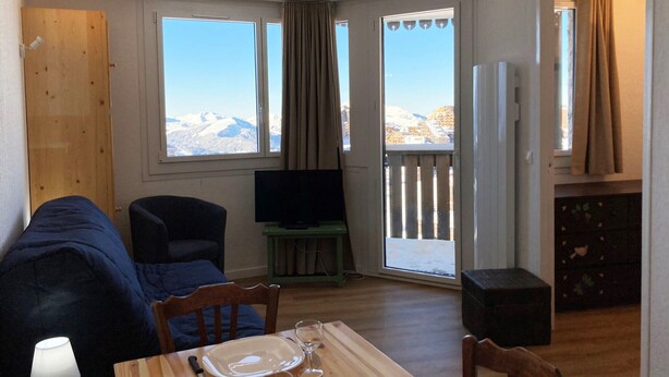 A cozy room with a sofa, a dining table with a plate and glasses, a TV on a stand, and a view of mountains through large windows.