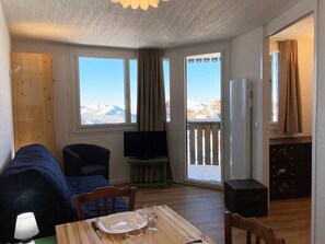 Une pièce chaleureuse avec un canapé, une table à manger avec une assiette et des verres, un téléviseur posé sur un meuble, et une vue sur les montagnes à travers de grandes fenêtres.