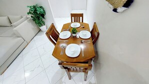 Un comedor con una mesa de madera dispuesta para cuatro personas, rodeada de sillas de madera. Se ve una planta en maceta a la izquierda y una cesta tejida colgada en la pared.