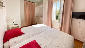 A bedroom with two beds, a TV, a bedside table with a lamp, a window with curtains, and a wardrobe. - Charming Holiday Home With Garden in the Cotentin (Fierville-les-Mines)