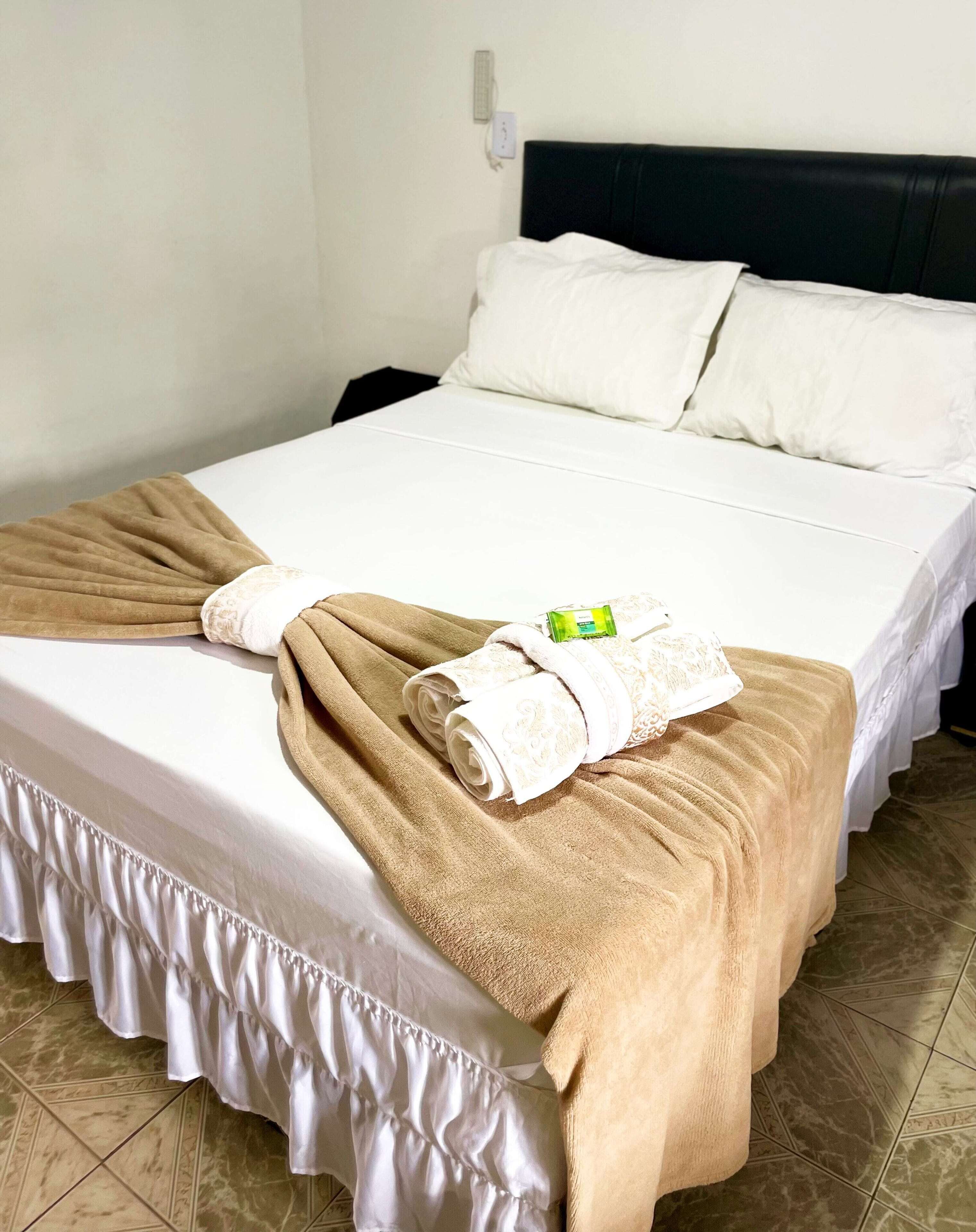 Una cama bien hecha con sábanas blancas, una manta color beige y una toalla enrollada.