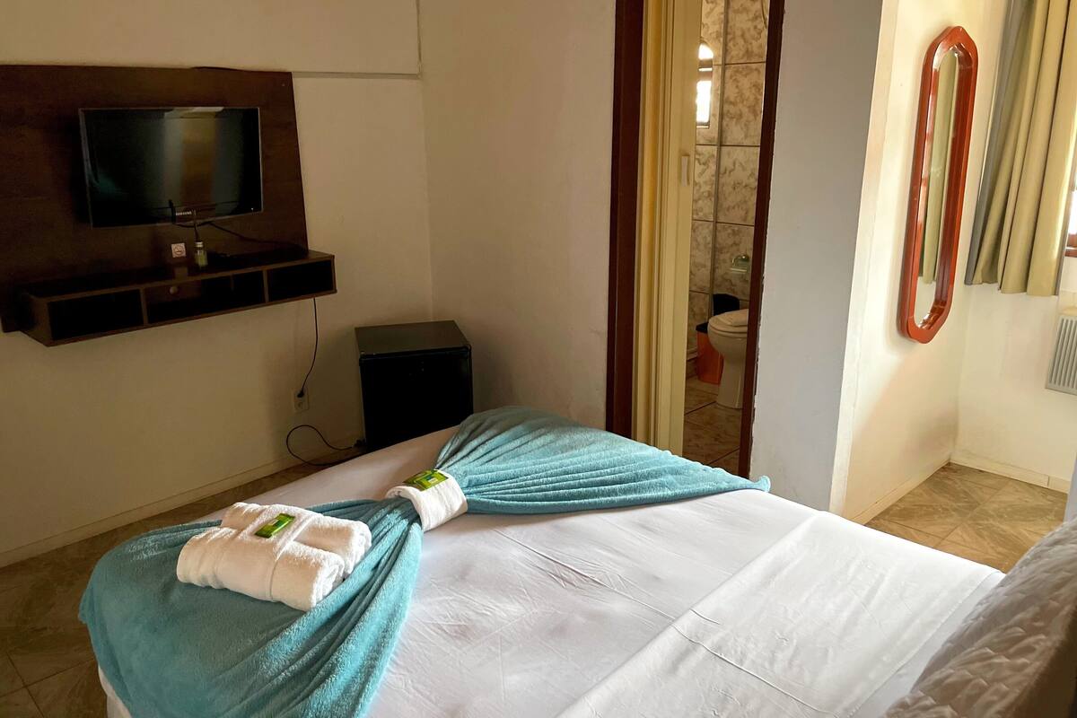A hotel room with a bed, a TV mounted on the wall, a small black cabinet, and a bathroom visible through an open door.