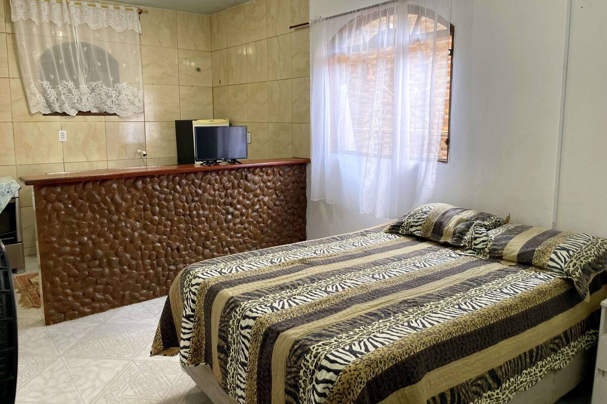 A bedroom with a bed, a TV on a wooden dresser, and a window with sheer curtains.