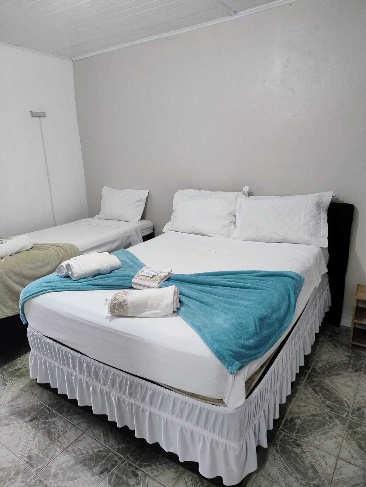 Una cama individual con ropa de cama blanca y una manta color turquesa, una mesita de noche pequeña y una lámpara fijada a la pared.