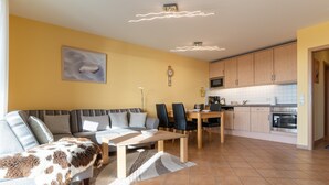 Ein modernes Wohnzimmer mit einer Couch, Sesseln, einem Couchtisch und einem Esstisch. Es gibt eine Küchenzeile mit Schränken und Geräten.