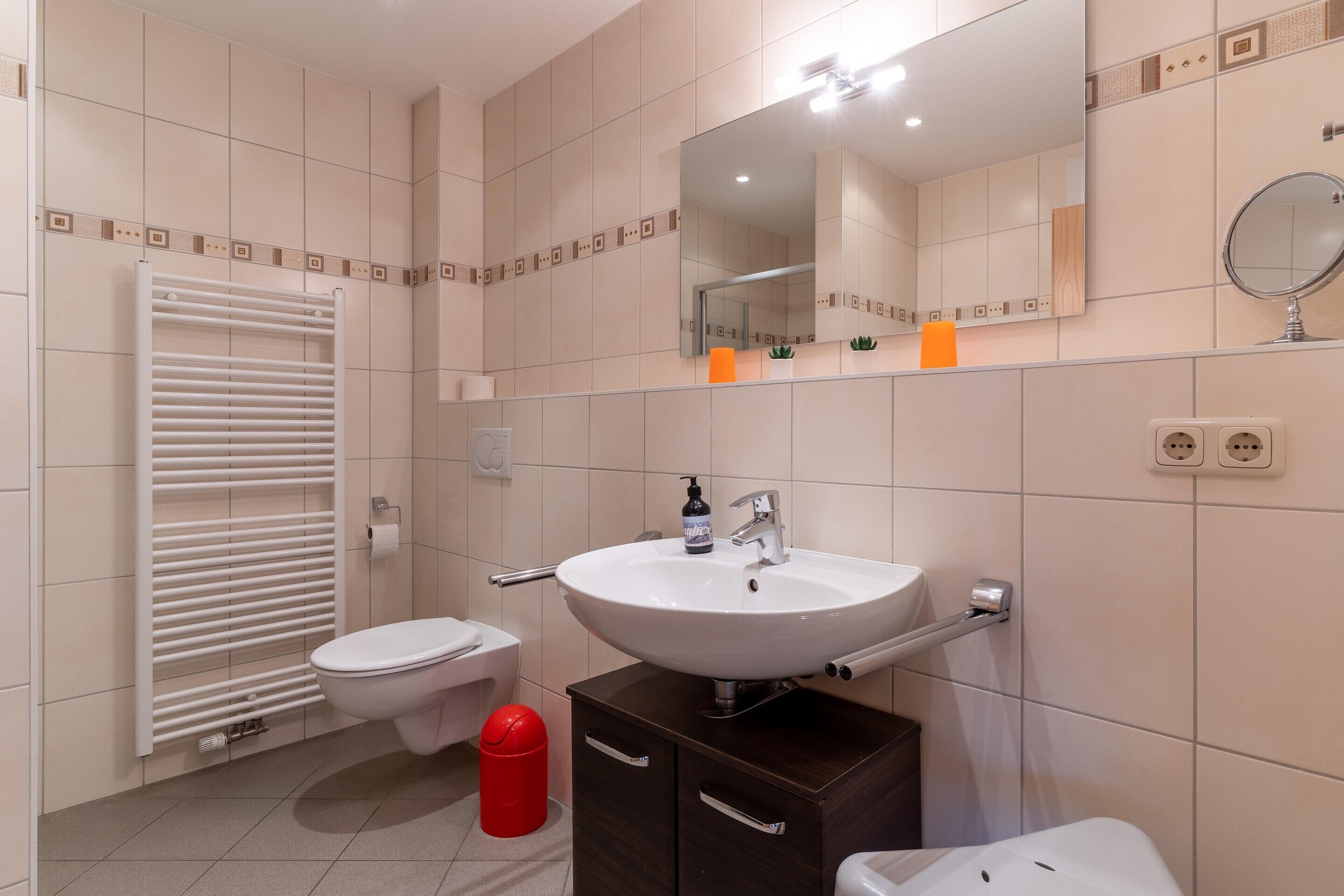 A modern bathroom with a white sink, a toilet, a red trash bin, and a towel warmer.