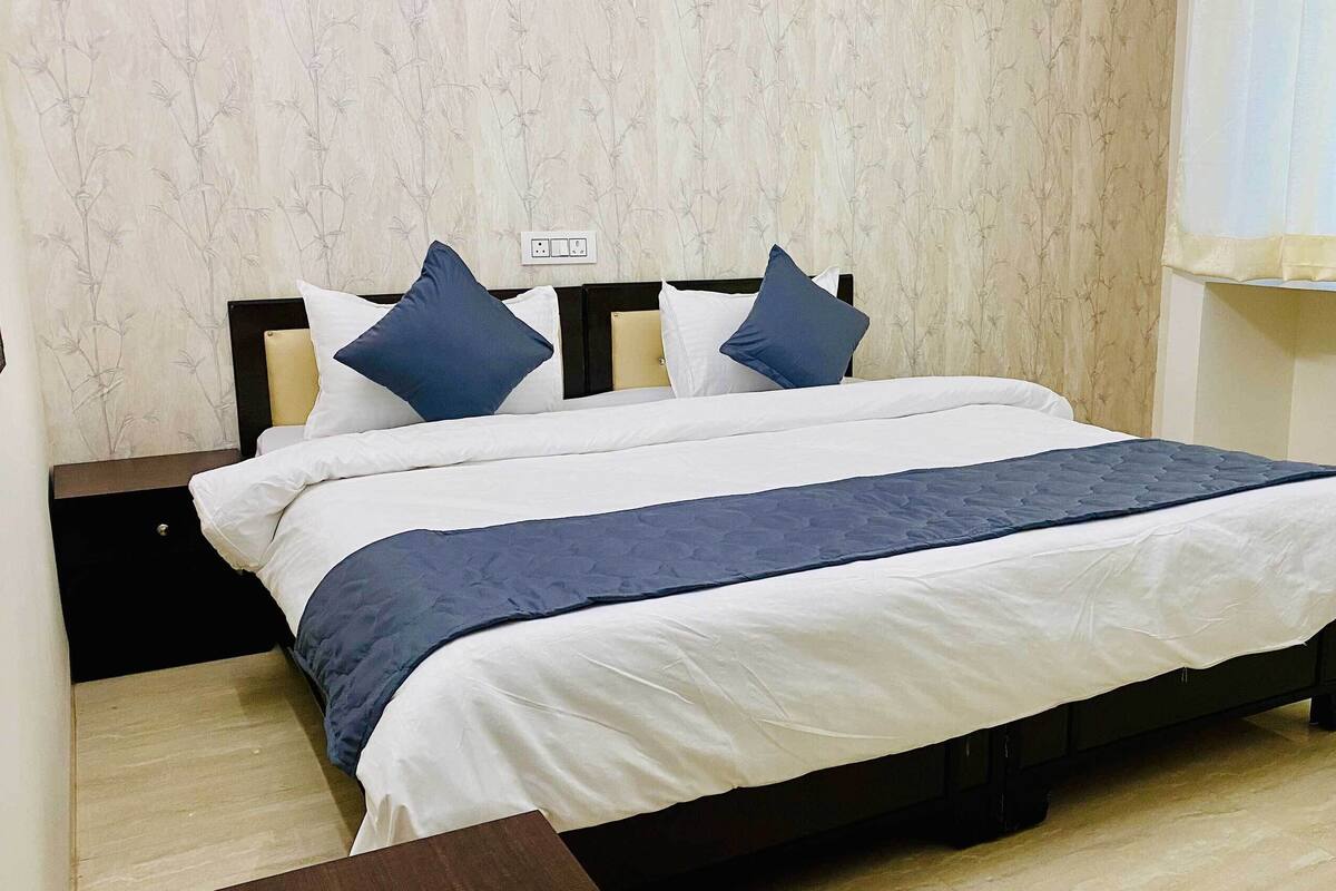 A neatly made bed with a dark blue bedspread and pillows, a wooden headboard, and a light-colored wall with a subtle pattern.