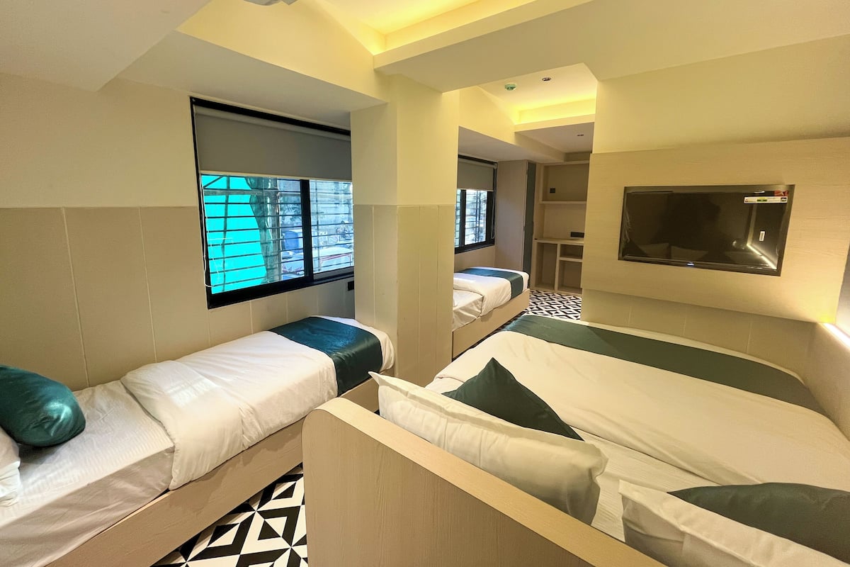 A hotel room with two single beds, a TV mounted on the wall, and a window with blinds.