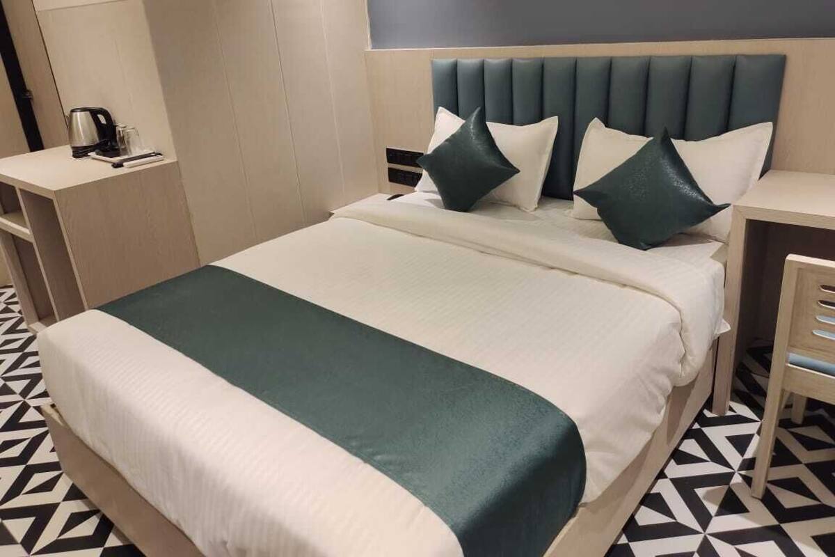 A neatly made bed with a green and white bedspread, a headboard with multiple pillows, a bedside table with a kettle, and a geometric patterned floor.