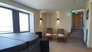 A room with a massage table, two massage chairs, a small table, and a wicker chair. There is a window with blinds, a door with an emergency exit sign, and a staircase leading to another floor. - Luxury Family Holiday Home With Wellness (Weiswampach)