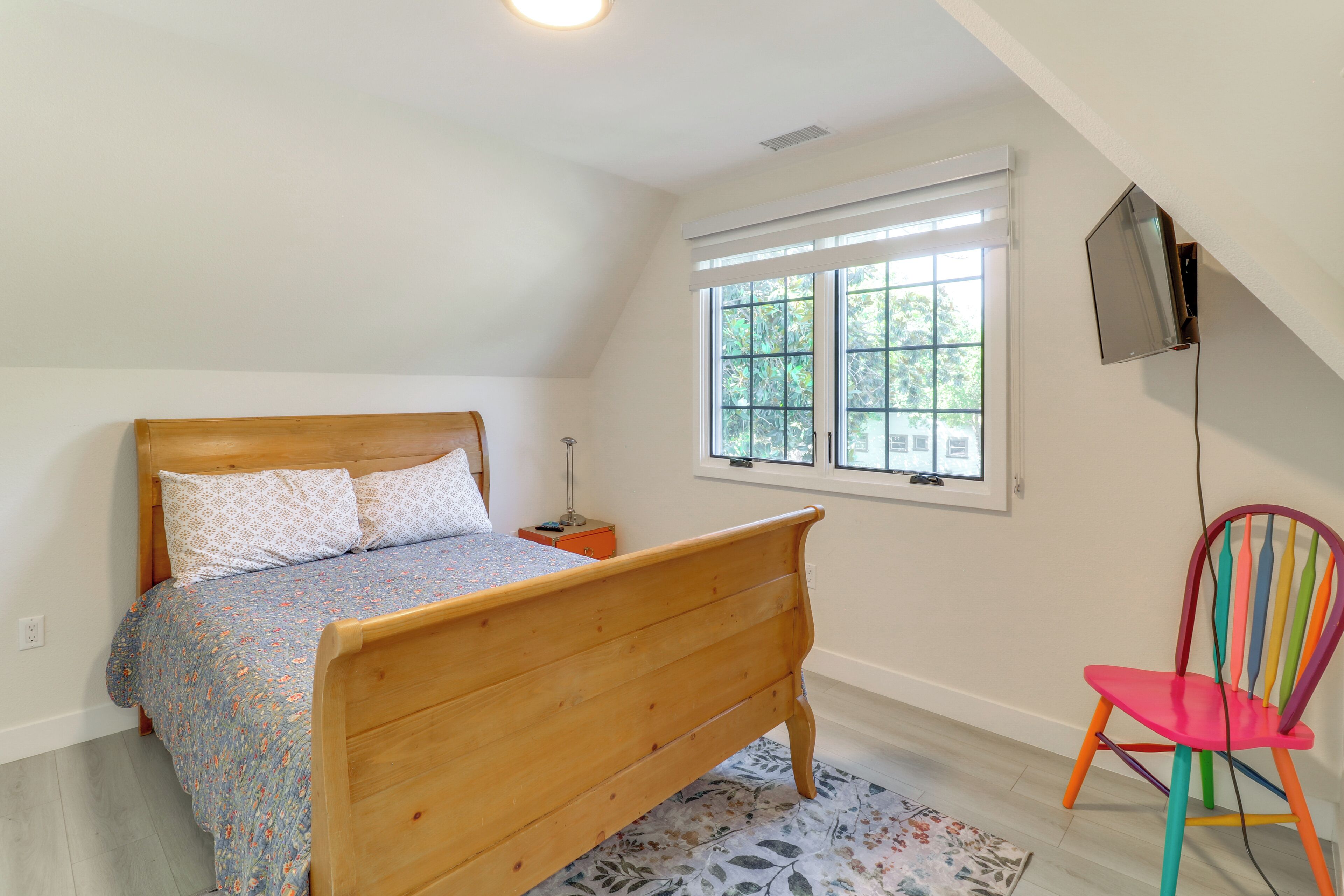 A bedroom with a wooden bed, a window with blinds, a colorful chair, and a TV mounted on the wall.