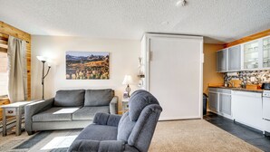 A modern living room with a grey sofa, a small table, and a lamp. There is a painting on the wall and a kitchen area with white cabinets and a black countertop. - 2 Mi to Granby Ranch: Mtn-view Studio w/ Perks (Granby)
