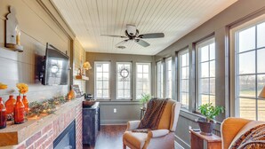 A cozy room with a fireplace, a ceiling fan, and large windows offering a view of the outdoors. - 3 Mi to Dtwn: Charming Farmhouse in Mount Joy! (Mount Joy)