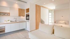A modern kitchen with white cabinetry, a built-in oven, and a sink. There is a wooden accent wall and a white sofa in the adjacent living area. - Aristi House in Olimpiada (Aristotelis)