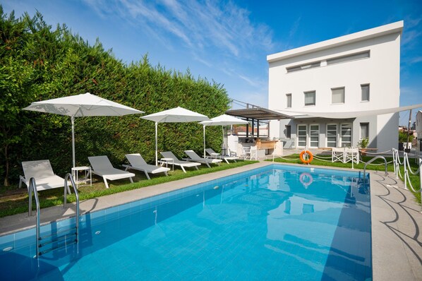 A swimming pool with lounge chairs and umbrellas, a modern building in the background, and a hedge. - Aristi House in Olimpiada (Aristotelis)