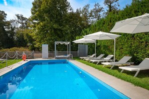 A rectangular swimming pool with a clear blue water surface, surrounded by a paved deck. There are white lounge chairs and umbrellas arranged for sunbathing, and a small covered seating area with a white railing. - Aristi House in Olimpiada (Aristotelis)