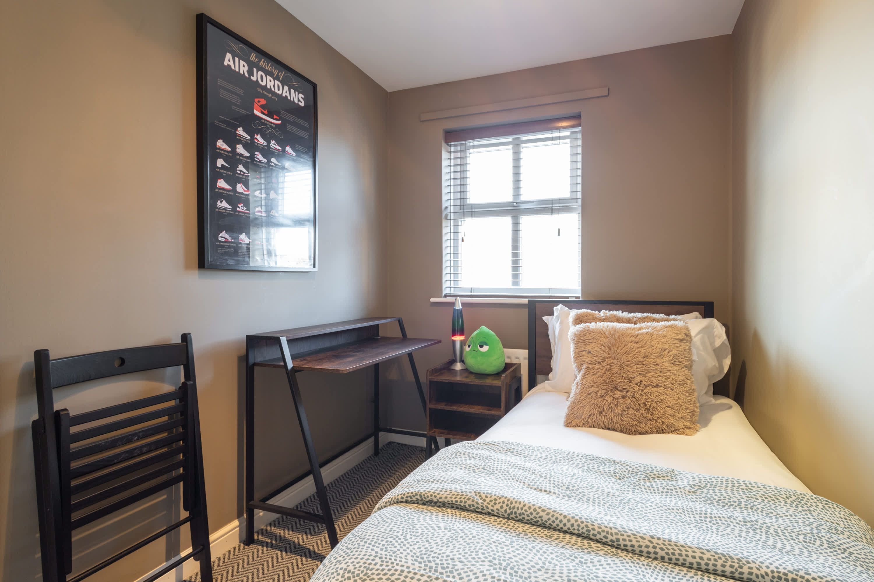 A bedroom with a bed, a desk, a chair, a window with blinds, and a poster on the wall.