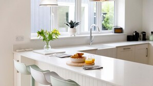 Une cuisine moderne avec un plan de travail blanc, deux tabourets de bar, un évier, une fenêtre avec des plantes et un coin petit-déjeuner avec des croissants et du jus d’orange.