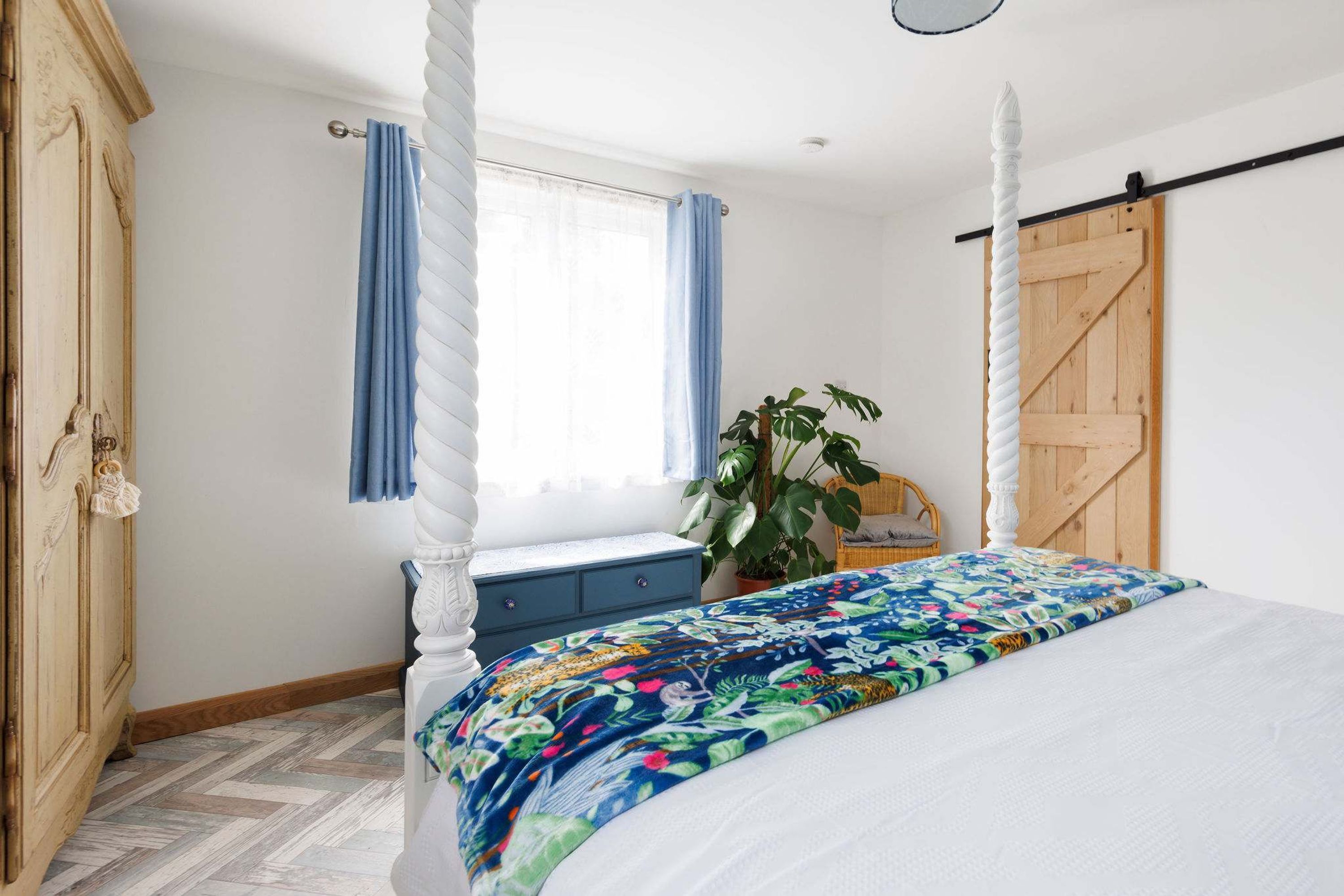 A bedroom with a four-poster bed, a wooden wardrobe, a window with blue curtains, and a potted plant.