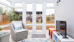 A sunlit room with a television, wicker furniture, and a view of a garden through glass doors.