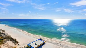 Vista aérea de una playa con un muelle que se adentra en el océano, arena y una estructura con techo azul.