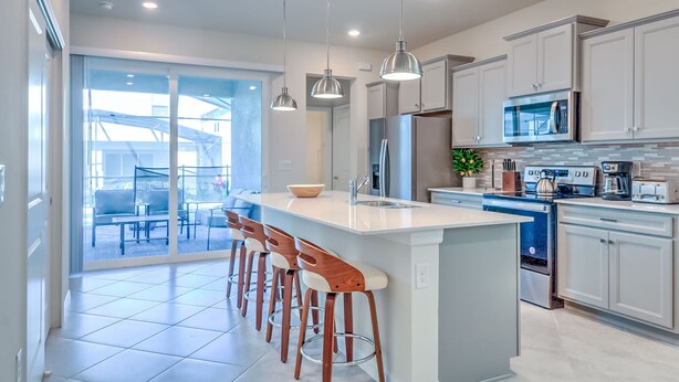 A modern kitchen with a central island, bar stools, stainless steel appliances, and a sliding glass door leading to an outdoor seating area. - 16235 Luxury Retreat Your Perfect Getaway Family Home (Clermont)