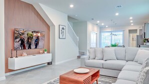 A modern living room with a large flat-screen TV, a wooden accent wall, a staircase, a grey sofa, and a kitchen area in the background. - 16235 Luxury Retreat Your Perfect Getaway Family Home (Clermont)