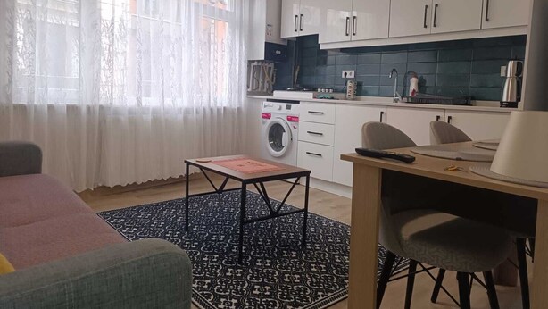 Un espace de vie compact comprenant un coin cuisine, un lave-linge, un coin repas avec des chaises et une grande fenêtre agrémentée de rideaux légers.