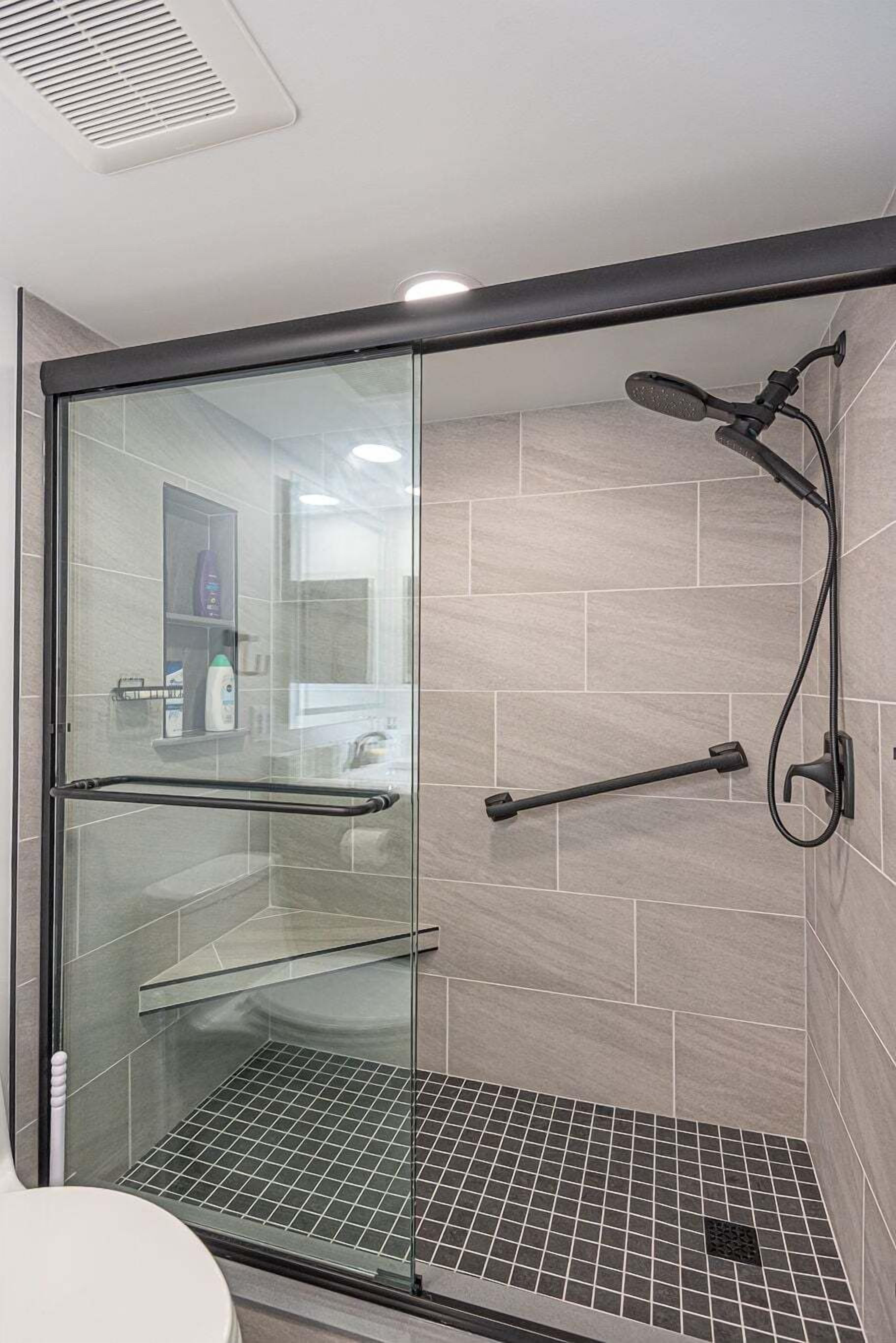 A glass-enclosed shower with a handheld showerhead, a glass shelf, and a metal grate floor.