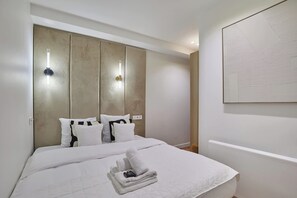 Ein modernes Hotelzimmer mit einem großen Bett, einer Kopfteilbeleuchtung, einem ordentlich bezogenen Bett mit Handtuch und Pflegeprodukten sowie einem großen abstrakten Gemälde an der Wand.
