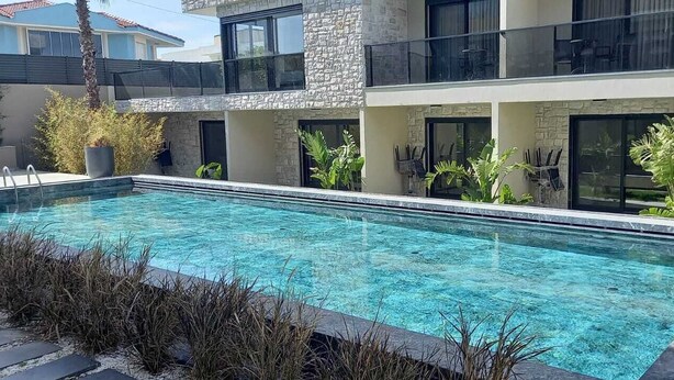 Una piscina rectangular con agua azul cristalina, rodeada por un edificio de piedra y vidrio con balcones y plantas en macetas.