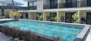 Une piscine rectangulaire à l’eau d’un bleu cristallin, entourée d’un bâtiment en pierre et en verre doté de balcons et agrémentée de plantes en pot.
