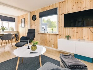 Una sala de estar moderna con una mesa de centro redonda blanca, dos sillones grises, un sofá negro, un mueble blanco y un televisor de pantalla plana montado en la pared.
