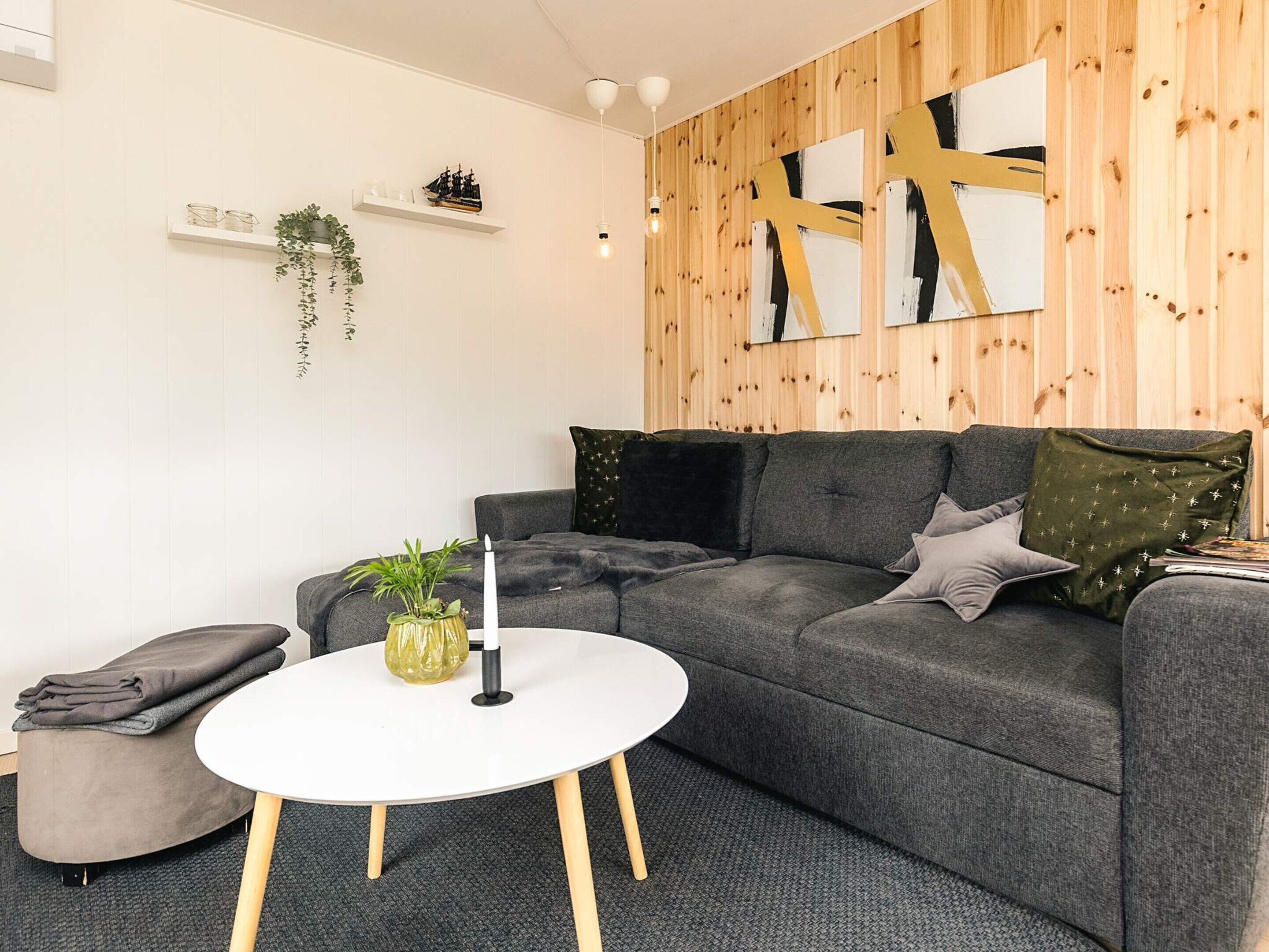 En moderne stue med en grå sofa, et hvidt sofabord og en lille grå skammel. Væggene er beklædt med træpaneler og prydet med abstrakt kunst samt reoler med dekorative genstande.