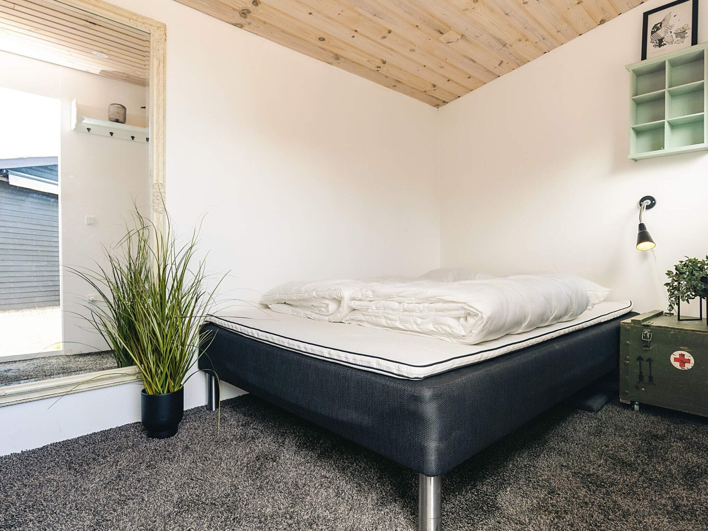 A bedroom with a bed, a potted plant, a nightstand, a wall-mounted shelf, and a window.