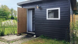 A small, dark wooden cabin with a single door and window, surrounded by a garden.