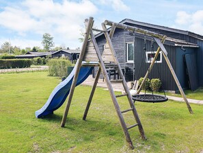A wooden swing set with a slide in a grassy yard, a small garden, and a dark wooden building in the background. - 6 Person Holiday Home in Hadsund (Hadsund)