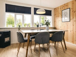 A dining area with a wooden table and chairs, large windows with blinds, a pendant light, and a framed picture on the wall. - 6 Person Holiday Home in Hadsund (Hadsund)