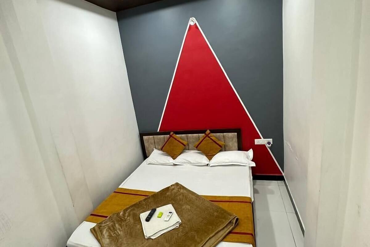 A compact hotel room with a bed, a small table with a brown cloth, a red triangular wall decoration, and a ceiling fan.