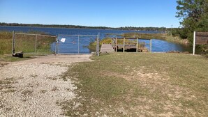 A gravel path leading to a wooden dock over a calm lake, surrounded by grassy areas and trees.