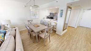A modern living room with a wooden dining table, white sofas, and a kitchen area with white cabinets and a microwave. - Emerald Towers 904 2 Bedroom Condo (Destin)