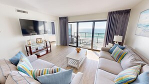 A modern living room with a large flat-screen TV, a sofa set with striped pillows, a coffee table, and a view of the ocean through large windows.