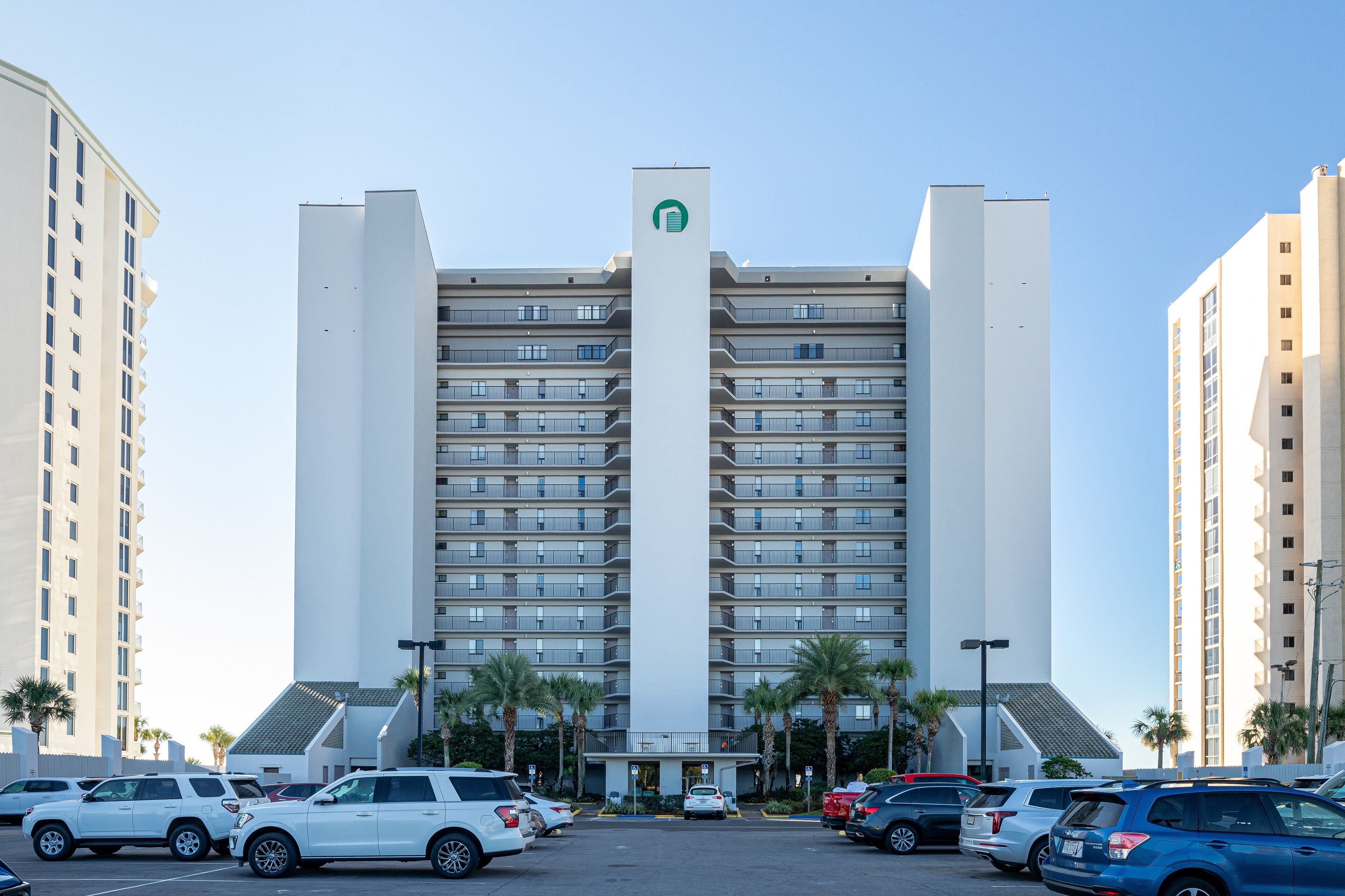 A multi-story hotel with a central vertical feature, parking lot, and surrounding palm trees.