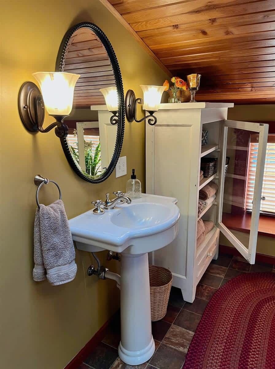 A bathroom with a pedestal sink, two wall-mounted lights, a mirror, and a cabinet with open doors revealing shelves.