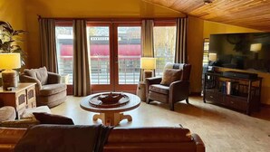 A cozy living room with a wooden ceiling, a television, and a large window with curtains. - Downtown Sequim Penthouse, Elegant Decor (Sequim)