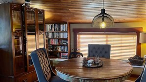 A round wooden dining table with a chandelier, surrounded by blue upholstered chairs. A large wooden bookcase filled with books is in the background. - Downtown Sequim Penthouse, Elegant Decor (Sequim)