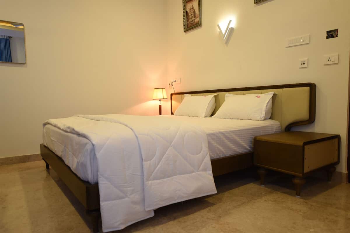 Una cama bien tendida con cubrecama y almohadas blancas, una mesita de noche con lámpara, y una lámpara de pared.