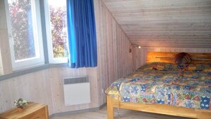 Une chambre avec un plafond en bois, un lit recouvert d’une couette aux couleurs vives, une table de chevet et une fenêtre ornée de rideaux bleus.